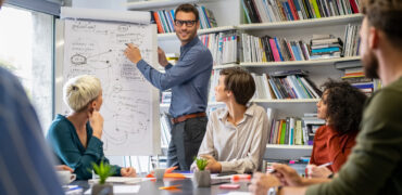 Confident businessman wearing spectacles and giving a presentation in office. Leader briefing his creative business team with annual goals. Mid adult business man training multiethnic businesspeople in conference room.