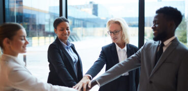 Four happy intercultural business people making pile of hands while standing in circle and looking at center